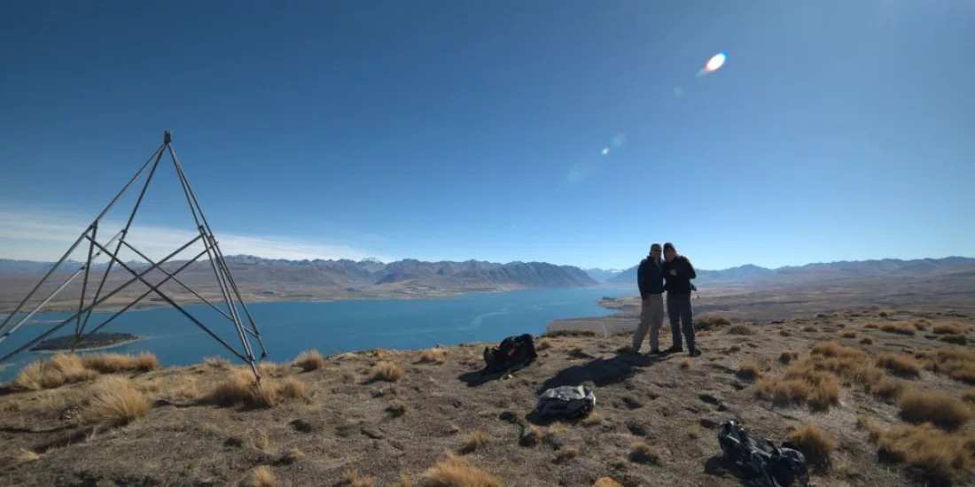 Two hikers posing for a photo near the trig station at the summit of Mount Hay with Lake Tekapo in the background