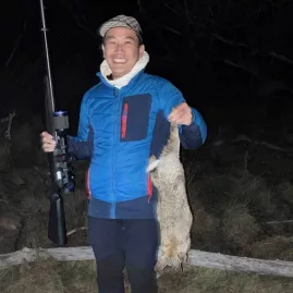 Smiling hunter holding a rabbit and rifle at night during a small game hunting experience near Lake Tekapo