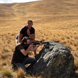 Guide assisting a guest with aiming a rifle during pest control training on Tekapo’s high country terrain