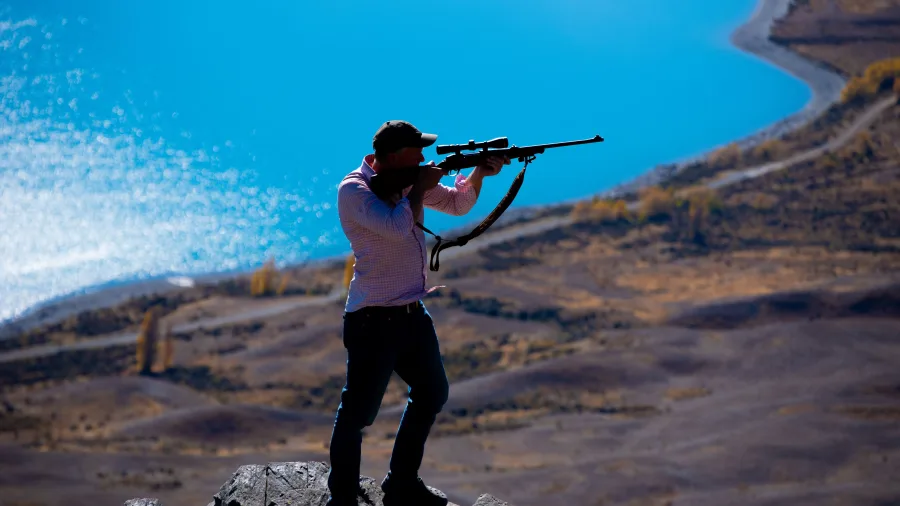 Hunter aiming a rifle on a rocky outcrop above Lake Tekapo during a guided small game hunting tour