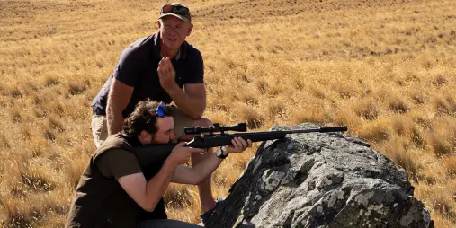 Guide assisting a guest with aiming a rifle during pest control training on Tekapo’s high country terrain