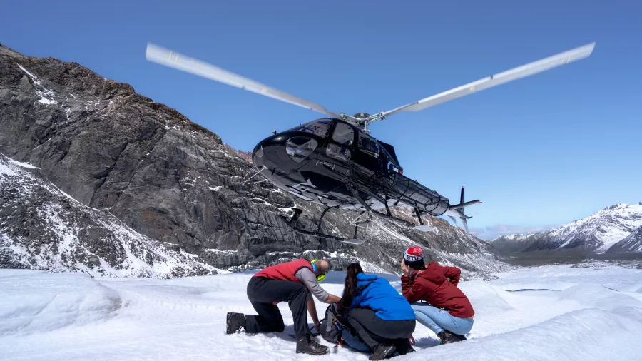 Helicopter landing on icy terrain while a group prepares gear beside it under clear skies