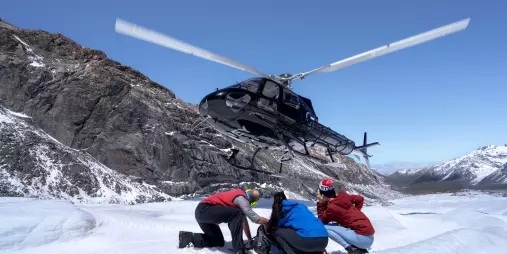 Helicopter landing on icy terrain while a group prepares gear beside it under clear skies