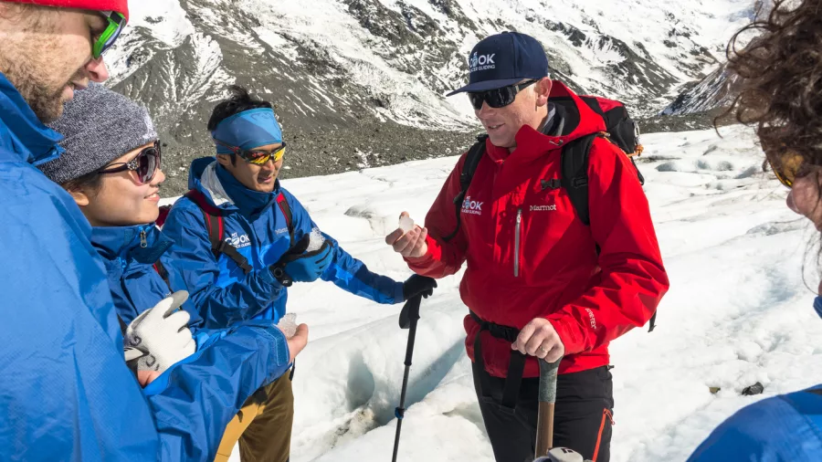 Glacier guide briefing a group of hikers on the ice with Mount Cook peaks in the background.