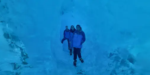 Three hikers standing in a narrow glacier tunnel with shimmering blue ice walls.