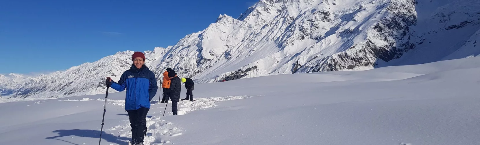 Snowshoer walking along a snowy trail with blue skies and mountain peaks in the background.