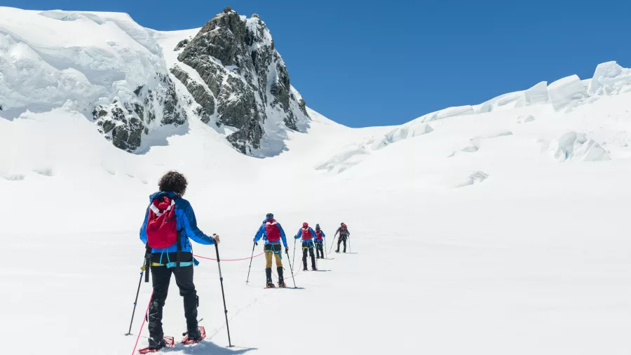 Roped snowshoers hiking across a wide glacier plateau beneath rugged mountain peaks.