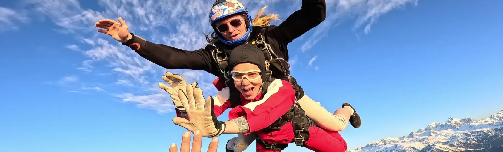 Tandem skydivers mid-air reaching for a high five with Mount Cook below
