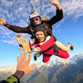 Tandem skydivers mid-air reaching for a high five with Mount Cook below