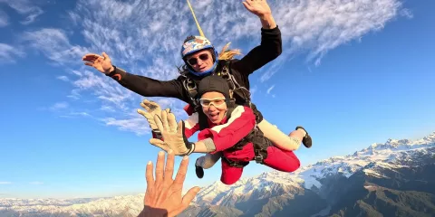 Tandem skydivers mid-air reaching for a high five with Mount Cook below