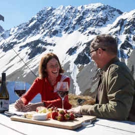 Couple enjoying wine and gourmet platter at an alpine picnic table with snow-covered mountains in the background