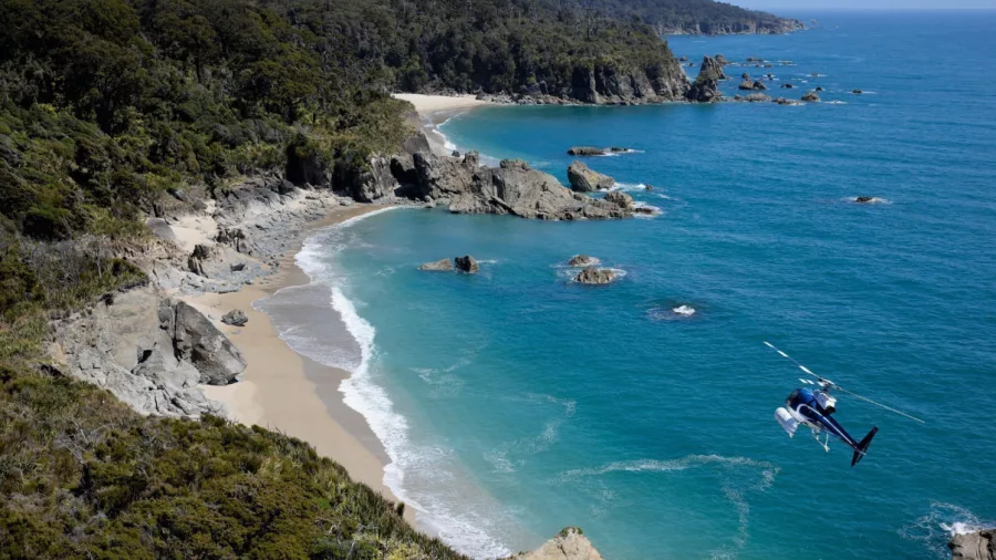 Helicopter flying above a rugged West Coast beach with turquoise waves and native forest