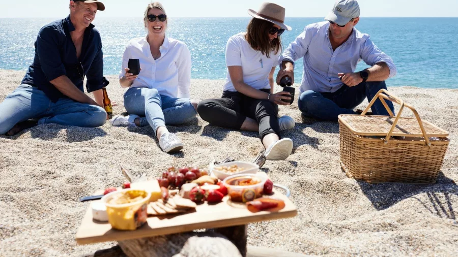 Group enjoying a gourmet platter on a pebbled West Coast beach with blue ocean in background