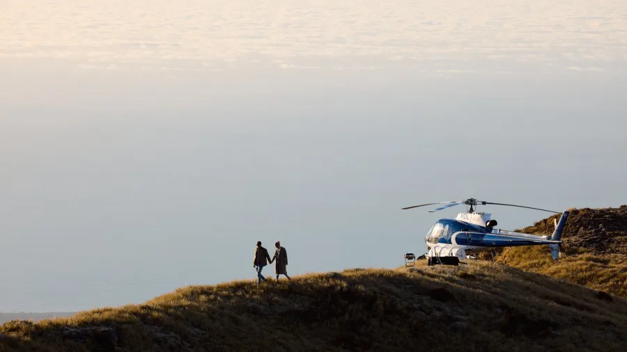 Couple walking across alpine ridge with private helicopter and vast ocean view