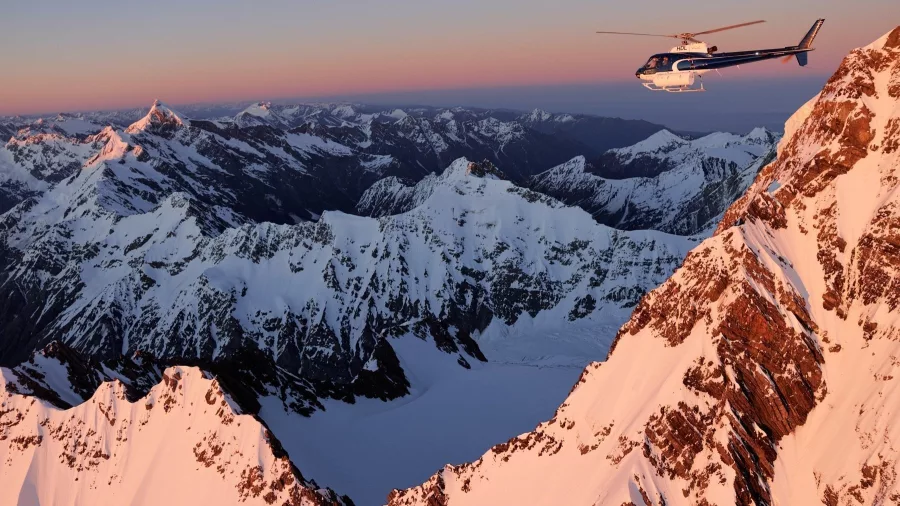 Helicopter flying past snow-covered ridge at sunset with sharp alpine peaks