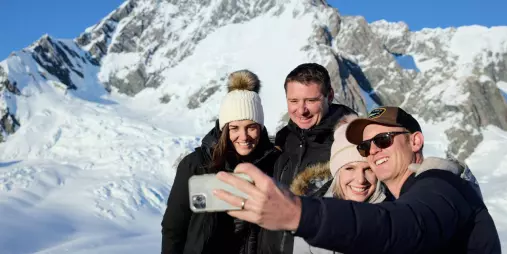 Group taking a selfie on the snow with Franz Josef Glacier and rugged peaks behind