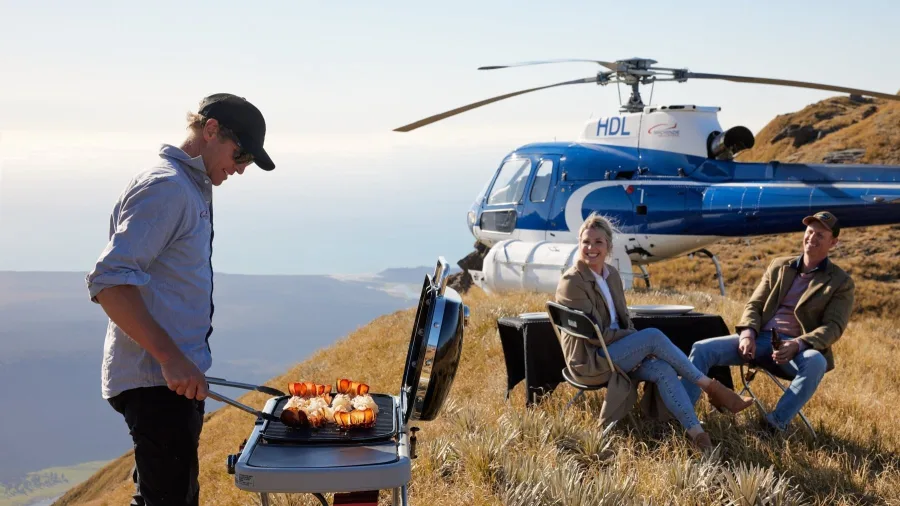 Private barbecue setup on a mountain peak with helicopter and couple enjoying coastal views
