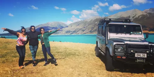 Group of travellers posing beside a 4WD vehicle with Lake Tekapo and Southern Alps in the background