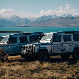Two 4WD Tekapo Adventures vehicles parked by the turquoise waters of Lake Tekapo with mountains in the background