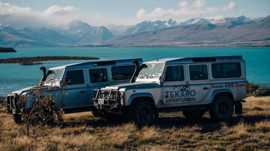 Two 4WD Tekapo Adventures vehicles parked by the turquoise waters of Lake Tekapo with mountains in the background