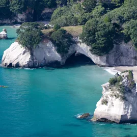 Aerial view of Cathedral Cove with kayakers paddling in turquoise waters near white limestone cliffs and sandy beach on the Coromandel Peninsula, New Zealand