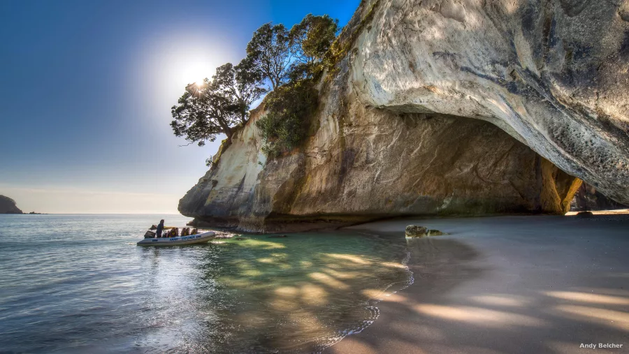 Tour boat arriving at Cathedral Cove beach beneath towering limestone cliffs and native forest on the Coromandel Peninsula, New Zealand