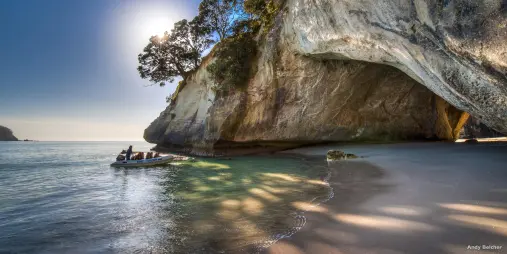 Tour boat arriving at Cathedral Cove beach beneath towering limestone cliffs and native forest on the Coromandel Peninsula, New Zealand