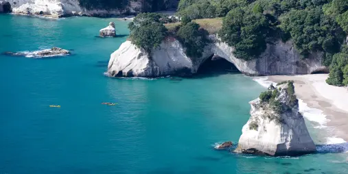 Aerial view of Cathedral Cove with kayakers paddling in turquoise waters near white limestone cliffs and sandy beach on the Coromandel Peninsula, New Zealand