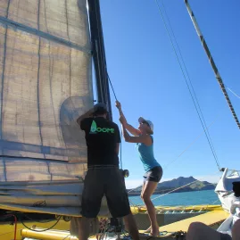 Boom Sailing crew and guest hoisting the sail aboard yellow catamaran on a sunny day