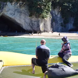 Two people relaxing on the catamaran looking toward Cathedral Cove’s beach and rock arch