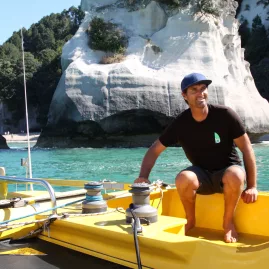Boom Sailing captain smiling at the helm with Cathedral Cove cliffs in the background