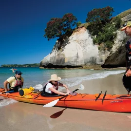 Guide helping paddlers launch an orange kayak from the beach at Cathedral Cove