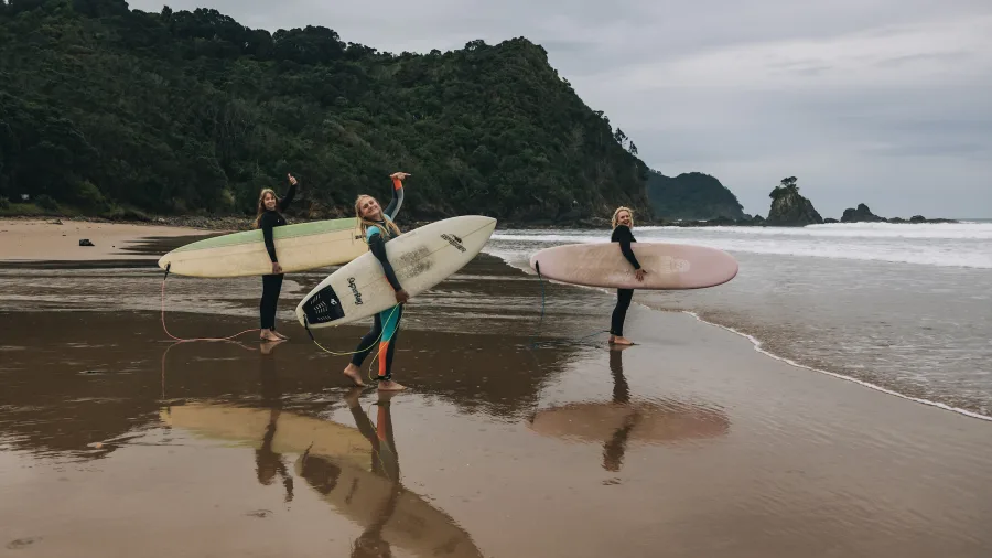 Group of women carrying surfboards on the wet sand at Sailors Grave Beach with waves and cliffs in the background on the Coromandel Peninsula, New Zealand.