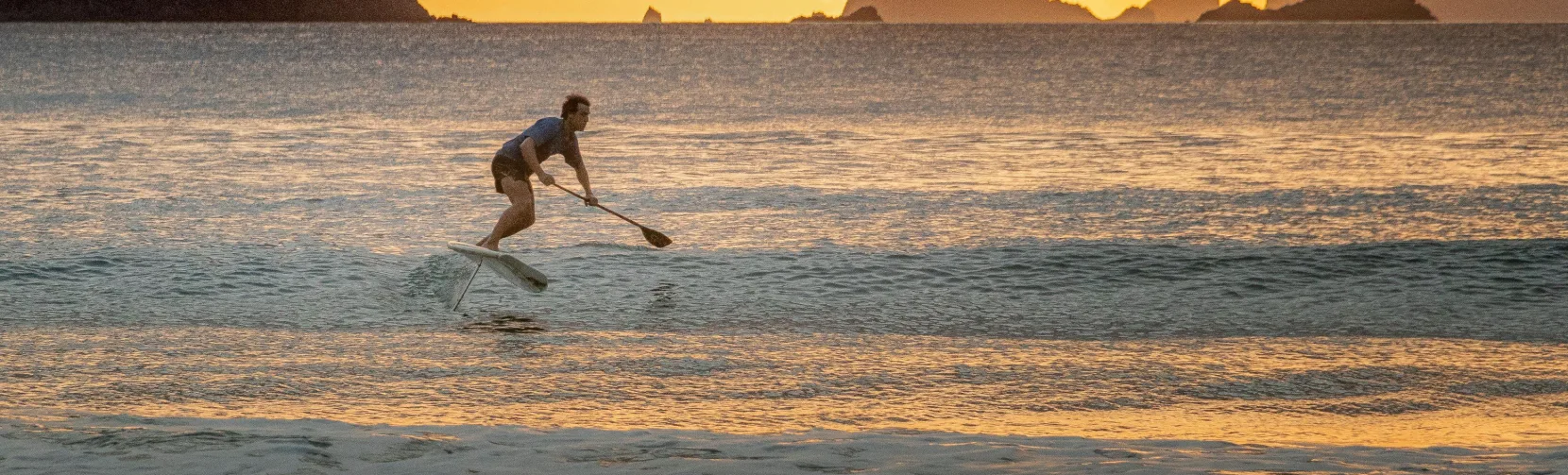Foil surfer riding the waves at sunset on Whitianga Beach with golden skies and offshore islands on the Coromandel Peninsula, New Zealand