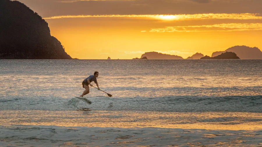 Foil surfer riding the waves at sunset on Whitianga Beach with golden skies and offshore islands on the Coromandel Peninsula, New Zealand