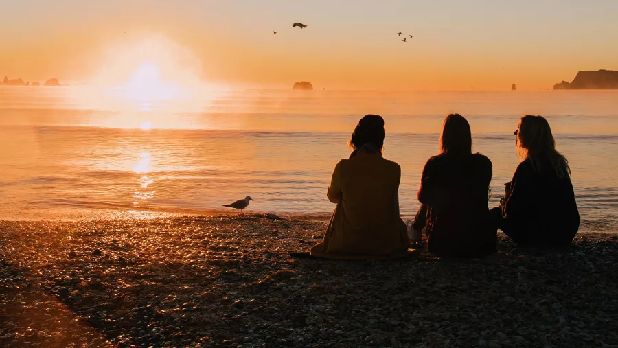 Three friends sitting on the sand watching the sunset over Whitianga Beach with seabirds flying above on the Coromandel Peninsula, New Zealand