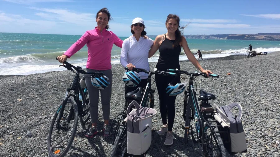 Three women with bikes on the shingle beach with Cape Kidnappers in the distance