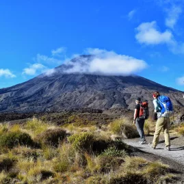 Three hikers walking a volcanic trail under blue skies with Mount Ngauruhoe in the background