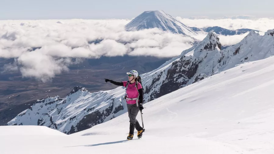 Female climber on snowy ridge of Mt Ruapehu pointing toward Mt Ngauruhoe across the clouds
