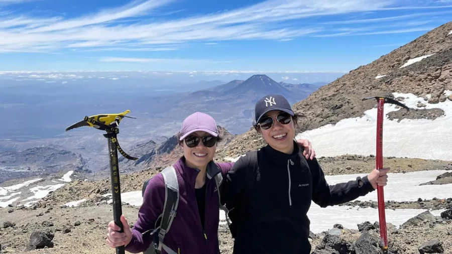 Two hikers smiling with ice axes on rocky terrain of Mt Ruapehu with volcanic peaks behind