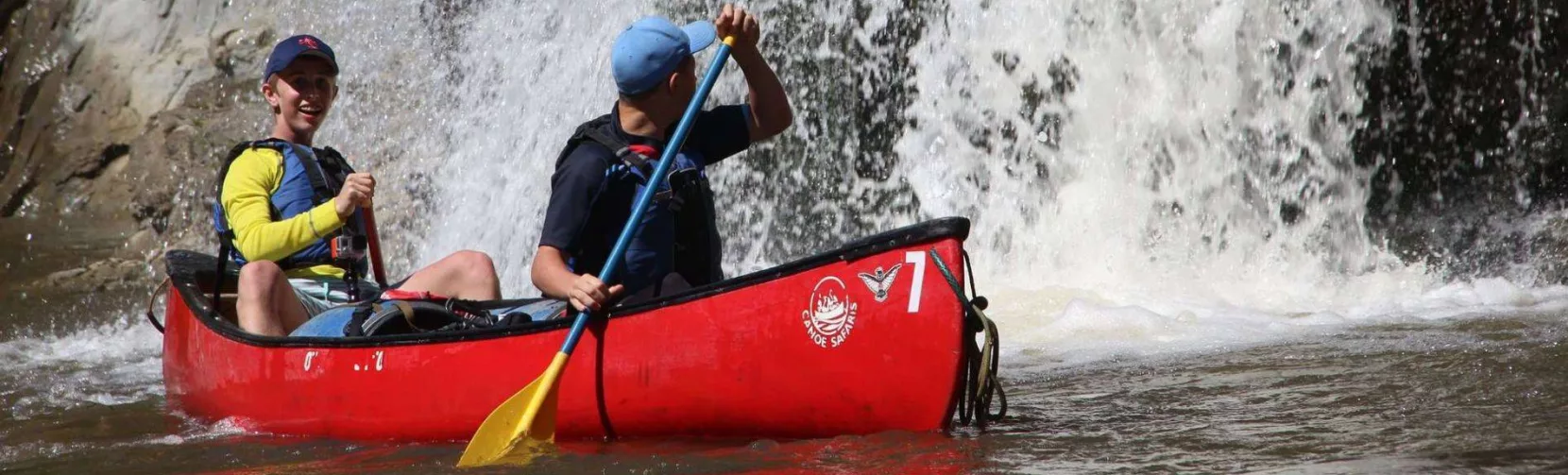 Two people in a red Canoe Safaris canoe smiling in front of a powerful waterfall on the Rangitikei River
