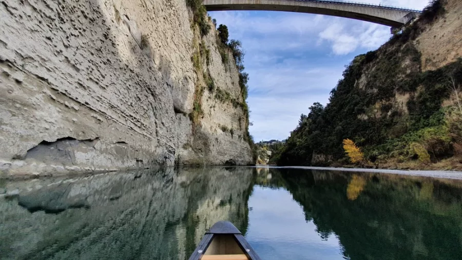 Canoe on calm water approaching the modern arched Utiku Bridge between towering papa cliffs on the Rangitikei River