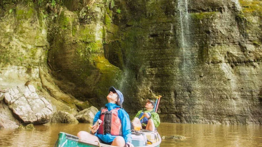 Two people in a green canoe gazing up at a small waterfall flowing down mossy cliffs on the Rangitikei River