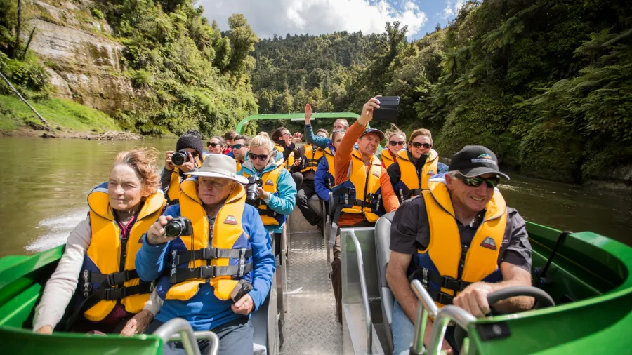 Jet boat passengers wearing life jackets, smiling, taking photos and enjoying the ride.