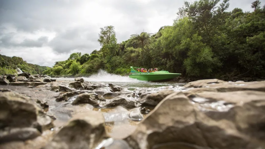 Green jet boat speeding along a shallow river with rocky banks in the foreground.