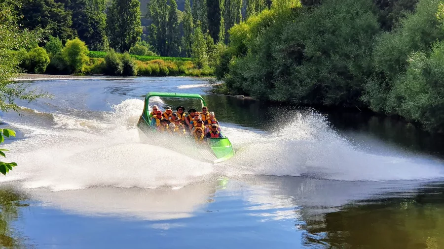 Jet boat creating a large water splash as it speeds along the calm, tree-lined Whanganui River.