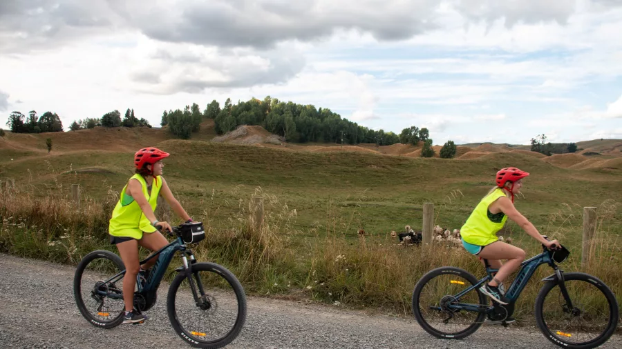 Two cyclists riding E-bikes along a gravel country road wearing hi-vis vests and helmets