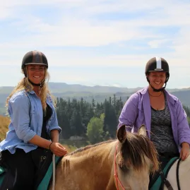 Two smiling women on horseback on a sunny hilltop trail ride with distant mountain views