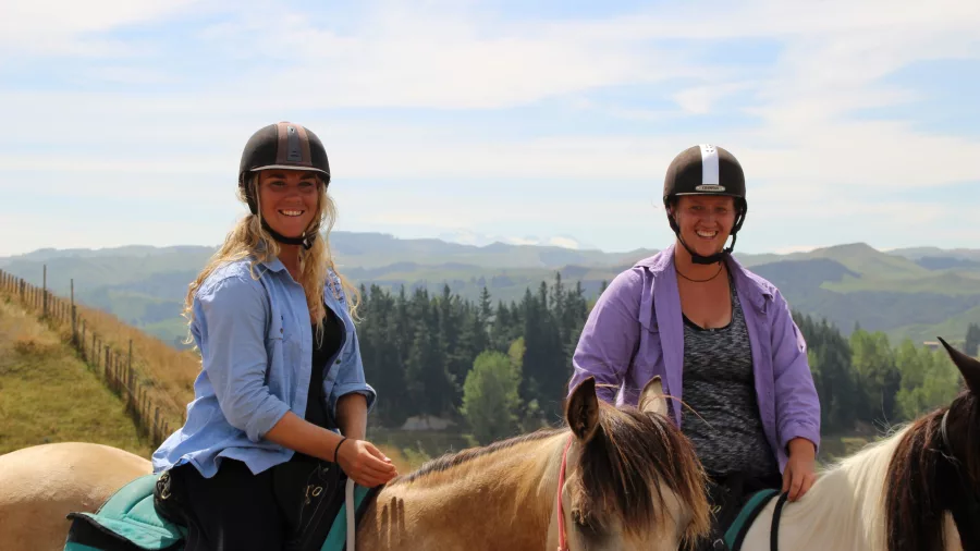 Two smiling women on horseback on a sunny hilltop trail ride with distant mountain views