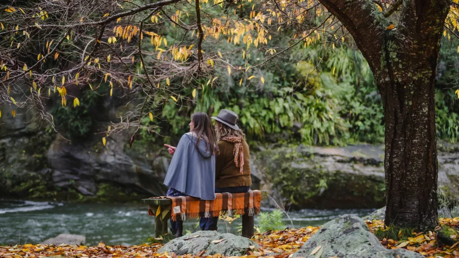Two people wrapped in blankets sitting on a bench beside the Rangitikei River in autumn